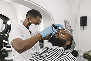 Barber wearing face mask and reusable gloves doing beard of customer in a barber shop.