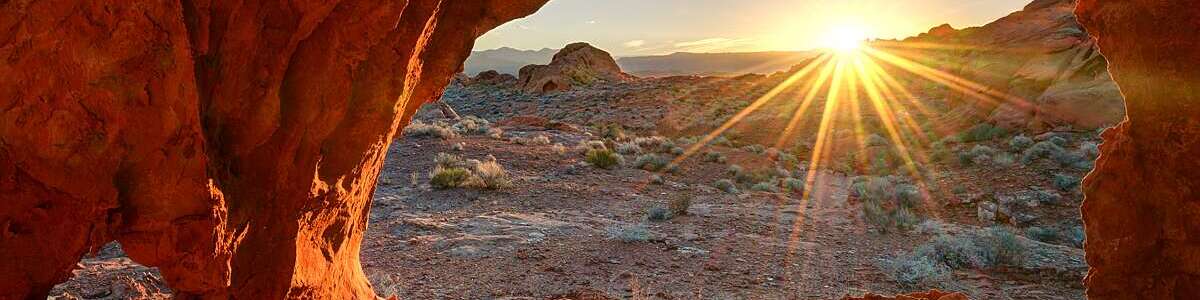 Dazzling ray of sun, seen through a red rock cavity, dances across a barren landscape at sunrise.