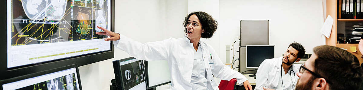 A clinical registrar reviewing a patients test results, looking at some information on a large monitor with some other doctors.