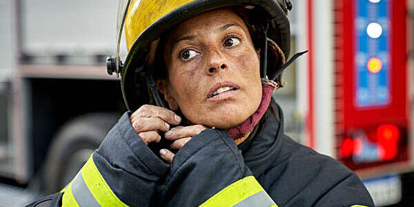 Head and shoulders close-up of late 30s Hispanic first responder with sooty face wearing turnout gear and securing helmet chin strap.